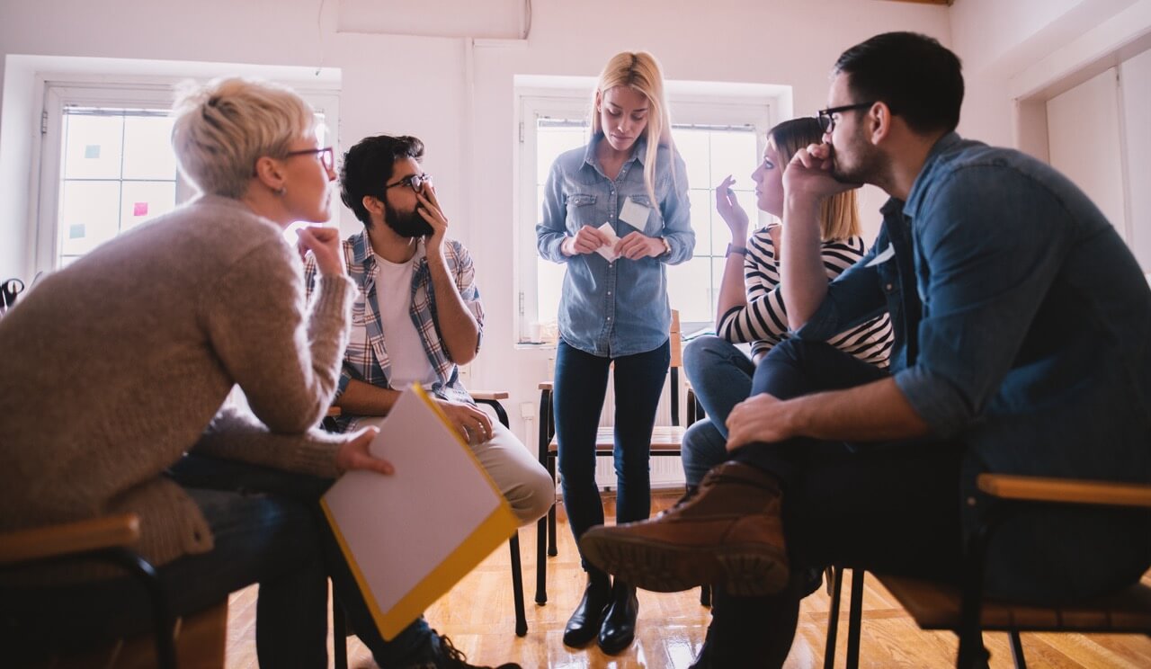 A group of people in a circle listening to a female speaking with a look of surprise on their faces