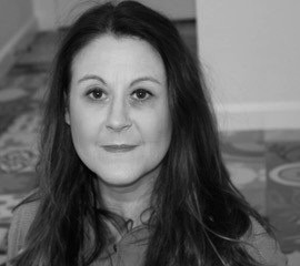Black-and-white portrait of Rachel Stephen, woman with long dark hair, looking directly at the camera with a neutral expression. She is indoors, seated against a patterned floor and plain wall background.