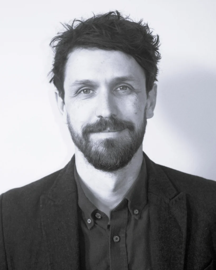 Black-and-white head-and-shoulders portrait of a David Sibley facing the camera. He has short dark hair and a beard and is wearing a dark jacket over a collared shirt. The image is closely cropped against a plain, light-coloured background with soft, even lighting.
