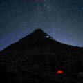 Dark mountain landscape beneath a star-filled night sky, with a small illuminated red tent in the foreground and the Milky Way visible overhead.