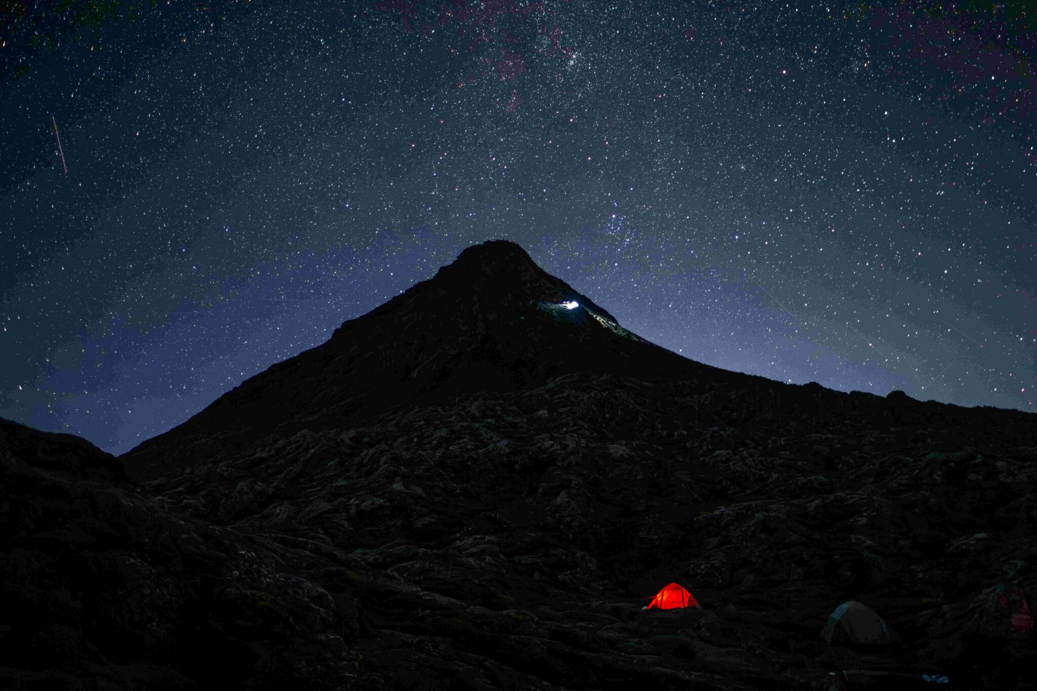 Dark mountain landscape beneath a star-filled night sky, with a small illuminated red tent in the foreground and the Milky Way visible overhead.