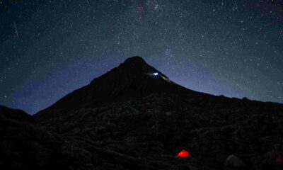 Dark mountain landscape beneath a star-filled night sky, with a small illuminated red tent in the foreground and the Milky Way visible overhead.
